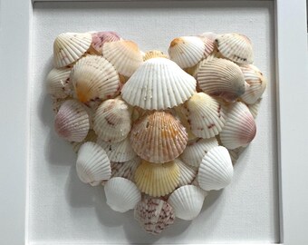 May include: A square white frame containing a heart-shaped arrangement of various seashells. The shells are primarily white, cream, and pale pink, featuring ribbed textures and fan-like shapes, mounted against a plain white fabric background.