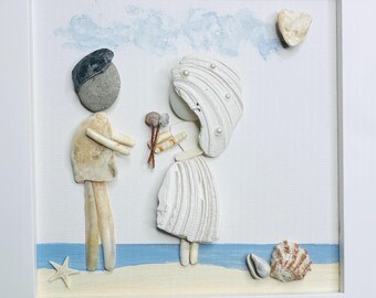 May include: A framed wall art piece featuring two figures crafted from seashells and stones. One figure wears a dark stone head, while the other has a white shell veil with small pearls. They hold a small bouquet of dried twigs on a painted beach.