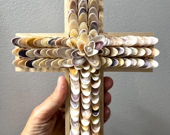 May include: A wooden cross decorated with layered, overlapping seashells in shades of cream, tan, and purple. The center of the cross features a flower-shaped arrangement of shells with a single small white pearl bead at the very center point.