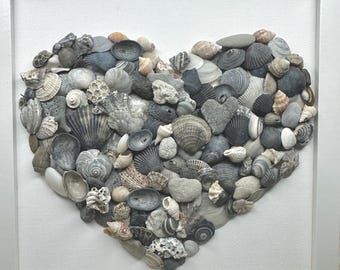May include: A square framed wall art piece featuring a collection of various gray, white, and tan seashells and small stones arranged in the shape of a heart on a white textured background. The initials DMAS are printed in the bottom left corner.