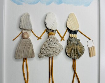 May include: A framed wall art piece featuring three figures crafted from seashells and twigs. The figures have shell hair, textured shell dresses, and twig limbs, standing on a painted blue line with a blue cloud and shell sun above.