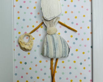 May include: A framed wall art piece featuring a figure constructed from seashells and twigs. The figure wears a skirt made of striped shells and holds a circular shell. The background is white with colorful polka dots. A single shell sits in the top right corner.