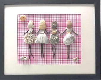 May include: A framed wall art piece featuring four figures crafted from various seashells and twigs. The figures are arranged in a row against a pink and white plaid background, with additional small seashells placed in the top left and bottom corners.