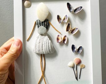May include: A framed seashell art piece featuring a figure made of shells, with a shell head, a gray and white striped shell dress, and brown twig arms and legs. Shell butterflies and flowers are also included in the artwork.