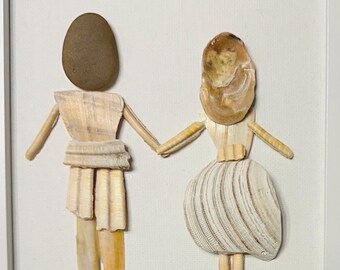 May include: A framed wall art piece featuring two figures made of seashells and a smooth brown stone, holding hands. Above them is a heart-shaped shell. The figures stand on a base of small, assorted pebbles and shells against a white background.