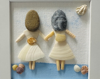 May include: A framed wall art piece featuring two figures crafted from seashells and stones. The figures have pebble heads, shell dresses, and shell limbs, holding hands against a white and blue background with additional shells at the base.