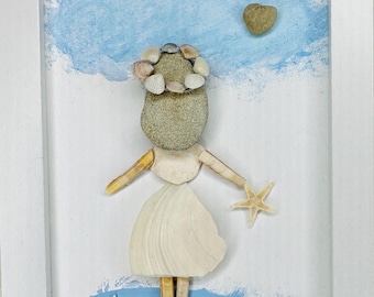 May include: A framed wall art piece featuring a figure constructed from seashells and stones. The figure has a stone head with a shell crown, shell arms, and a shell skirt. A starfish is held in one hand, set against a painted blue sky and sea.