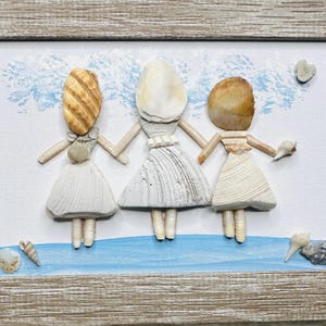 May include: A framed wall art piece featuring three figures crafted from various seashells. The figures have shell heads and bodies wearing white shell dresses, standing on a painted blue base against a white background with blue accents.