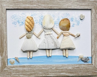 May include: A framed wall art piece featuring three figures crafted from various seashells. The figures have shell heads and bodies wearing white shell dresses, standing on a painted blue base against a white background with blue accents.