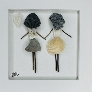 May include: A framed artwork featuring two figures crafted from seashells. One figure has a black shell hat and gray skirt, the other has a gray rock head and a beige shell skirt. They are holding hands with twig arms and legs.