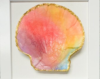 May include: A seashell with a multi-colored watercolor finish in shades of pink, orange, yellow, and blue, featuring a textured surface and a metallic gold painted edge, displayed inside a square white frame against a white background.