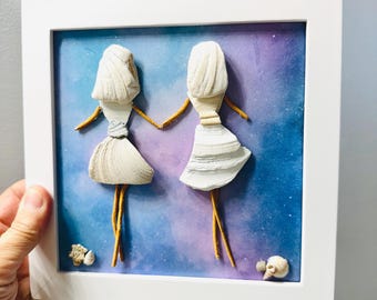 May include: A framed wall art piece featuring two figures crafted from white seashells and thin brown twigs. The figures are holding hands against a blue and purple watercolor background with small white dots. Two small seashells sit at the bottom.