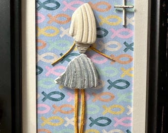 May include: A framed wall art piece featuring a figure constructed from two textured white seashells, with yellow string arms and legs, set against a light blue background patterned with small, multicolored fish icons and a small white cross.