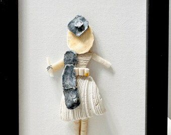 May include: A framed wall art piece featuring a figure constructed from various seashells. The figure wears a white shell dress, a dark gray shell hat, and a dark gray shell accessory, with a small white shell held in one hand against a white background.