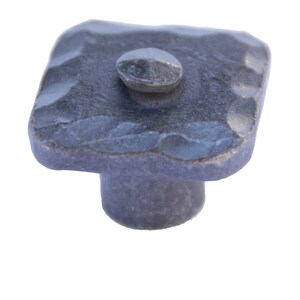 May include: A square-shaped, hammered metal cabinet knob with a round, slightly raised knob on top. The knob is a dark gray color.