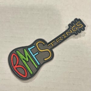 May include: Embroidered patch in the shape of a guitar. The guitar is gray with the words "BMFS" in red, blue, green, and yellow, and "STRINGS" in yellow. The patch has a black border.