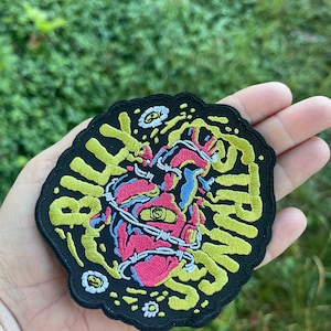 May include: Embroidered patch featuring a stylized heart pierced with barbed wire, with an eye in the center. The heart is pink, with blue and yellow accents. The patch has the word "STRINGS" in yellow, with a black border.
