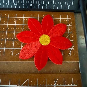 Può includere: Un fiore rosso ricamato con sei petali e un centro giallo. Il fiore è posto su una superficie marrone con un motivo a griglia e un righello lungo il bordo superiore. Il fiore misura circa 7,6 cm di diametro.