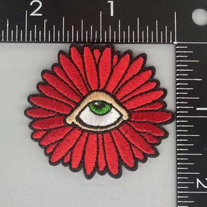 Puede incluir: Parche de flor roja bordada con un ojo blanco y un iris verde. El ojo está rodeado por un anillo beige.