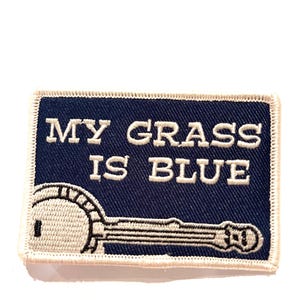以下が含まれることがあります： 白いステッチで刺繍された青いパッチ。パッチにはバンジョーと「MY GRASS IS BLUE」の文字が描かれています。