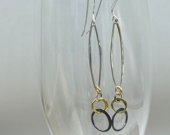 Op de afbeelding: Zilveren oorhangers met een lang, dun, ovaal ontwerp. De oorhangers hebben drie kleine cirkels aan de onderkant, één gouden en twee zwarte.