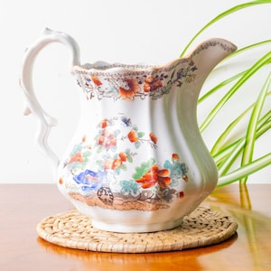 Może przedstawiać: Biały dzbanek z porcelany z kwiatowym wzorem w odcieniach pomarańczowego, czerwonego, niebieskiego i zielonego. Dzbanek ma żebrowane ciało i zakrzywiony uchwyt. Stoi na plecionym wiklinowym podkładce.