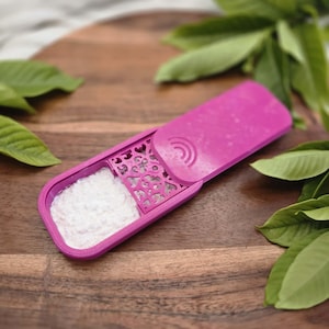 May include: A magenta plastic handheld sifter featuring a rectangular compartment filled with white powder, a decorative laser-cut metal grate with a heart pattern, and a sliding cover with a semi-circular embossed detail on the handle.