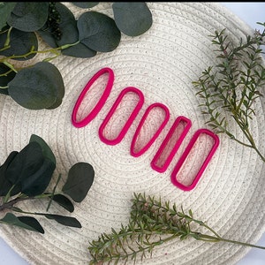 May include: A set of five bright pink oval-shaped cookie cutters. The cutters are arranged in a row, decreasing in size. The cutters are made of plastic and are on a cream-colored woven surface, with green foliage around them.