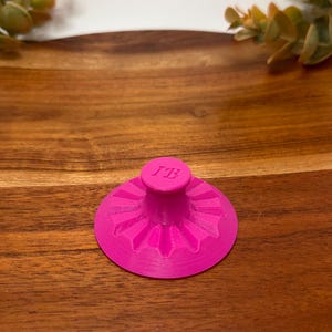 May include: A bright pink circular stamp with a central handle and a sunburst pattern radiating from the middle. The top of the handle features the embossed letters JB. The object has a flat base and a textured surface with visible 3D printed lines.