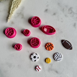 Pode incluir: Uma coleção de cortadores de biscoitos e detalhes decorativos com tema esportivo. O conjunto inclui cortadores de plástico rosa em forma de basquete, vôlei, futebol americano e beisebol. Também existem detalhes decorativos de basquete, bola de futebol, bola de tênis e vôlei.