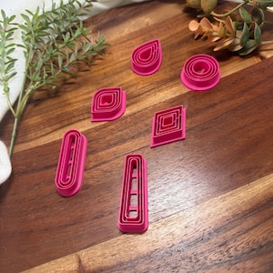 Könnte beinhalten: Ein Set aus sechs leuchtend pinkfarbenen Kunststoff-Ausstechformen in verschiedenen geometrischen Formen, darunter ein Tropfen, ein Kreis, eine Raute, ein Oval, ein Trapez und ein abgerundetes Rechteck, mit ineinander verschachtelten Konturen.