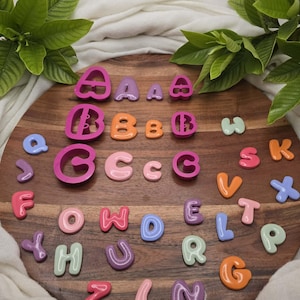 May include: A collection of multi-colored plastic alphabet letter cookie cutters and small, smooth, rounded letter charms in various shades of pink, purple, orange, blue, and light green, arranged on a circular wooden board with a visible grain.