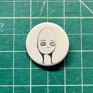 May include: A white button with a hand-drawn illustration of an egg-shaped figure with eyes and legs. The figure has a smiling face and is drawn in black ink.
