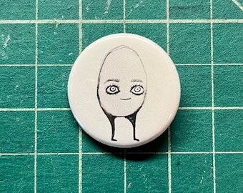 May include: A white button with a hand-drawn illustration of an egg-shaped figure with eyes and legs. The figure has a smiling face and is drawn in black ink.