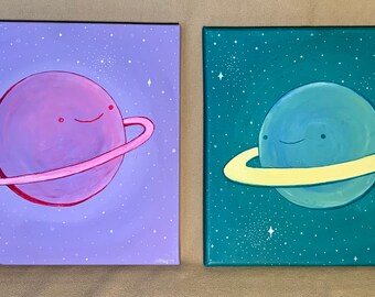 May include: Two paintings of planets with rings. The first painting is on a purple background with a pink planet and a pink ring. The second painting is on a teal background with a blue planet and a yellow ring. Both planets have a smiling face.
