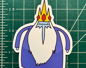 May include: A die-cut sticker featuring the Ice King from Adventure Time. The sticker depicts the character with a white beard, a blue robe, and a yellow crown with red gems. The sticker is on a green cutting mat.