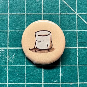 May include: A round button with a cartoon illustration of a white marshmallow with a smiling face and brown arms and legs. The marshmallow is sitting on a beige background.