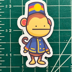 May include: A cartoon monkey wearing a blue uniform with yellow accents and a red and yellow hat with a yellow circle on top. The monkey has a brown face and a brown tail.