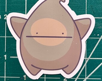 May include: A die-cut sticker featuring a cartoonish, blob-like character with a neutral expression. The character is primarily light brown with a lighter tan belly and darker brown accents. The sticker has a white border.