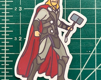 May include: A sticker featuring a cartoon illustration of Thor, a superhero character. Thor is depicted in a heroic pose, wearing a gray and black suit with a red cape, and holding his hammer. The sticker has a white border.