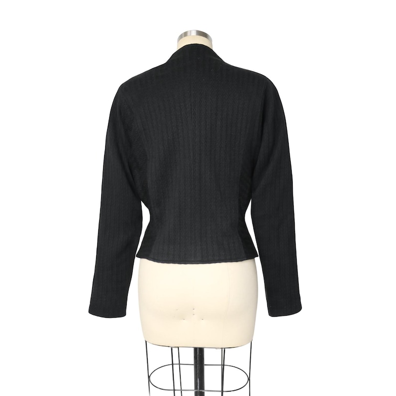 May include: Black, long-sleeved jacket with a textured, vertical stripe pattern. The jacket has a high, rounded collar and a fitted waist. The garment is displayed on a mannequin against a white background.