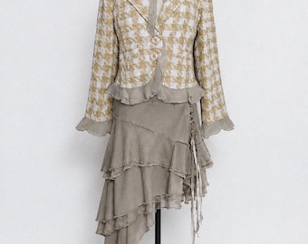 May include: A two-piece outfit on a mannequin. The jacket is a houndstooth pattern in gold and white, with a ruffled hem. The skirt is a layered, asymmetrical design in a muted gray-brown color. A fabric flower adorns the neckline.