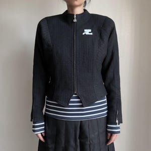 May include: Black zip-up jacket with a textured weave. The jacket features a high collar, a white logo on the chest, and zippers on the sleeves. The person is wearing a striped shirt underneath.