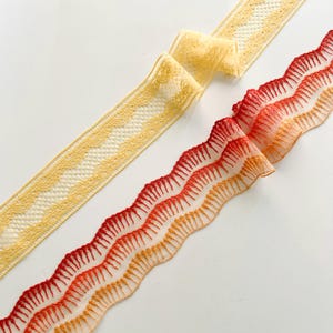 May include: Two decorative ribbons. One is a pale yellow with a scalloped edge and a delicate lace pattern. The other is a vibrant gradient of red and orange, also with a scalloped edge, creating a textured, layered effect. Both are on a white background.