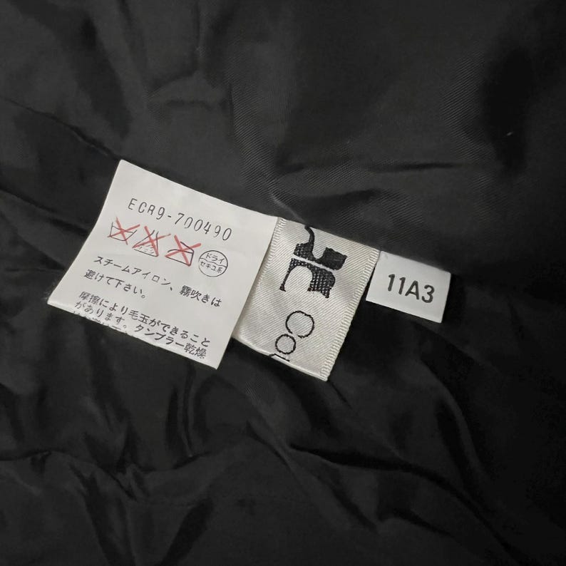 May include: Close-up of a black garment with attached tags. The tags display care symbols, the text "ECR9-700490", and the number "11A3". One tag has the word "Co" printed on it. The background is a dark, textured fabric.