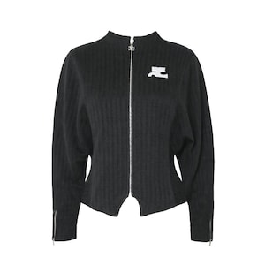 May include: A black zip-up jacket with a high collar and a white logo on the chest. The jacket has a textured, ribbed pattern and silver zippers on the front and sleeves. The sleeves are long and the jacket has a unique cut at the bottom.