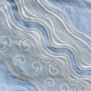 Puede incluir: Primer plano de un ribete de encaje blanco con un diseño ondulado delicado. El encaje presenta patrones intrincados y un borde festoneado, sobre un fondo de tela azul claro. El ribete incluye detalles arremolinados en forma de volutas.