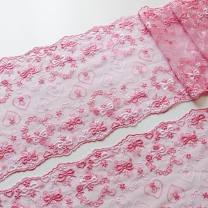 May include: Pink lace ribbon with embroidered hearts, bows, and floral details. The delicate trim is suitable for crafting and sewing projects. The ribbon is rolled up, showcasing the intricate design. The ribbon is measured in centimeters.