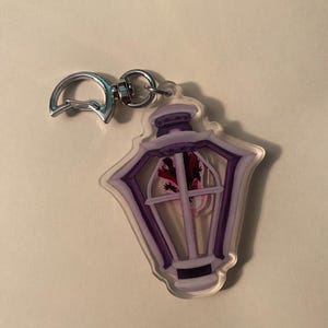 Puede incluir: Un llavero acrílico transparente con un diseño de linterna morada y blanca y un ajolote en el interior. El llavero tiene un cierre plateado para sujetarlo. La linterna tiene un diseño detallado con una ventana y una parte superior decorativa.