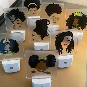 May include: Eight night lights with different images of Black women with various hairstyles, including afros, braids, and buns. Each night light has a white base and a clear glass front.